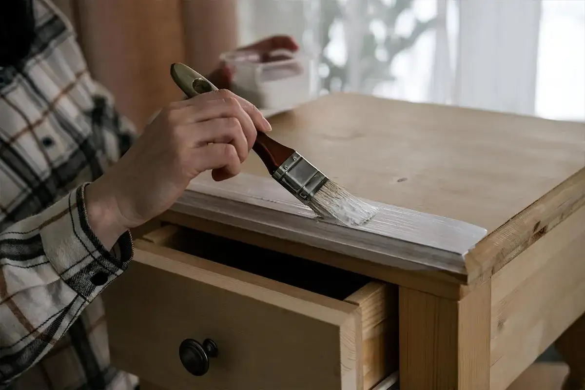 Persona pintando muebles de madera sin lijar, aplicando una capa de pintura blanca con brocha a un mueble de pino.